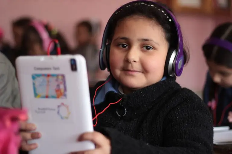 A girl with headphones holding a tablet in a classroom looks at the camera.