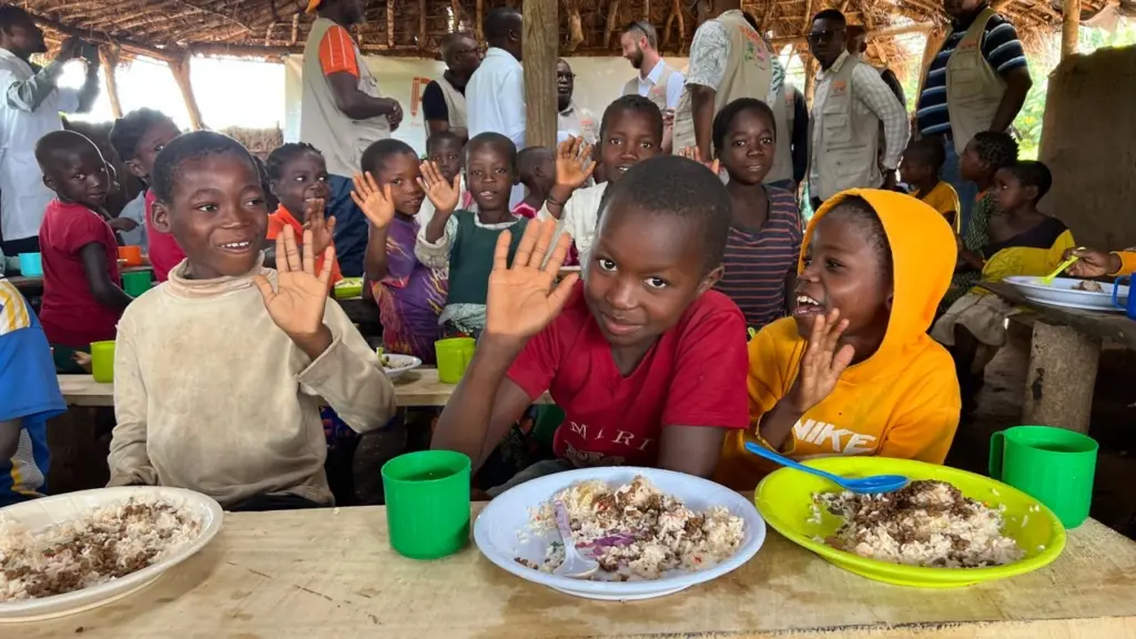 Smiling children with plates of food in front of them, some waving at the camera